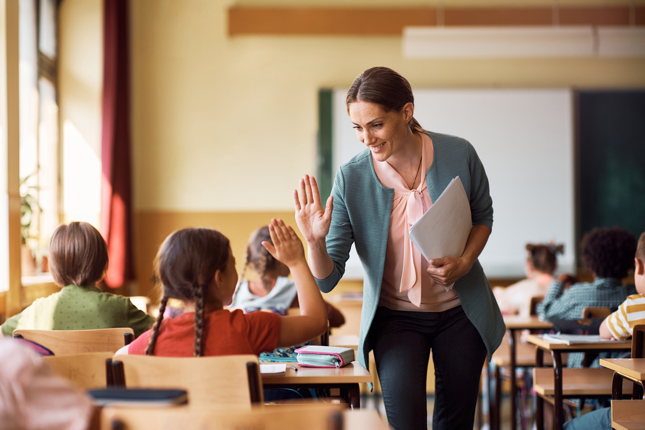 A teacher with classroom supplies in one hand, high-fiving a student in class