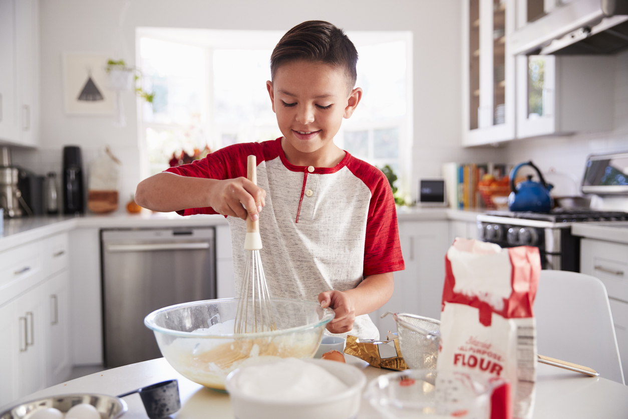 A young child in a kitchen mixing dough in a bowl with various cooking elements on the table
around him such as flour, eggs, and sugar