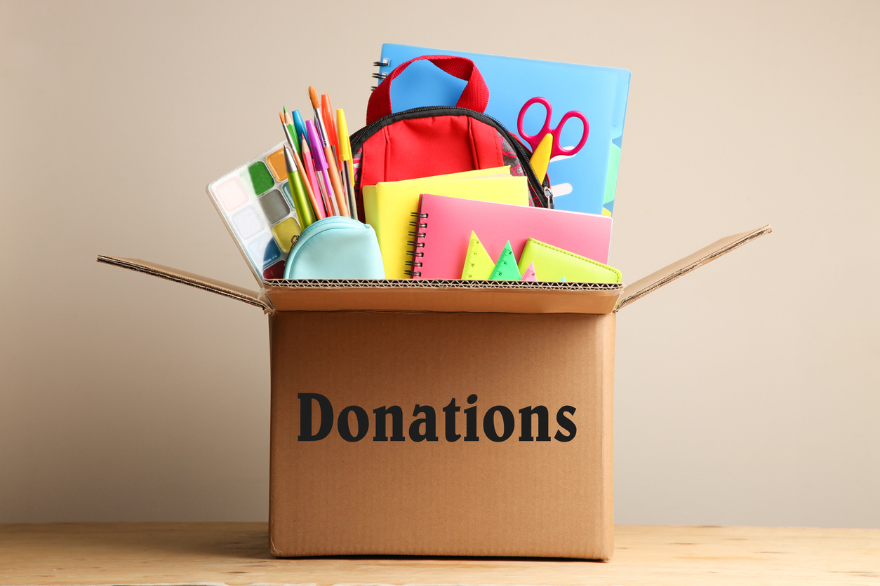 A box marked for donations filled with various school supplies like scissors, notebooks, pens, paints, and a backpack